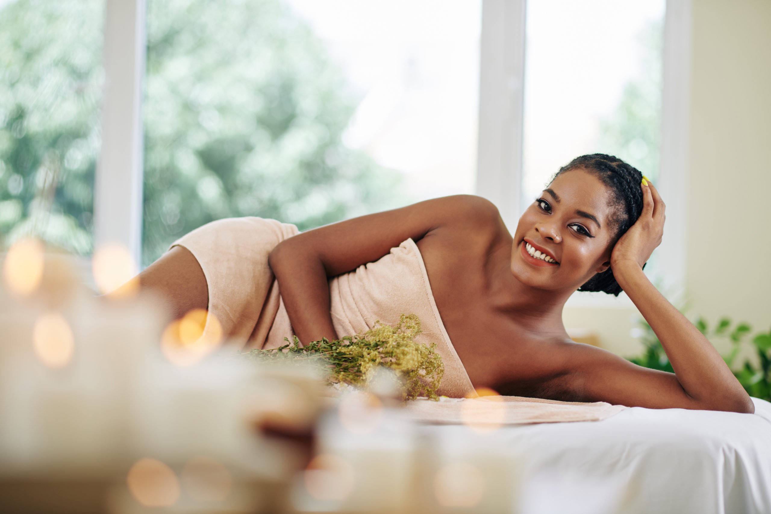 Portrait of happy smiling young Black woman relaxing on bed in spa salon and waiting for the next spa procedure