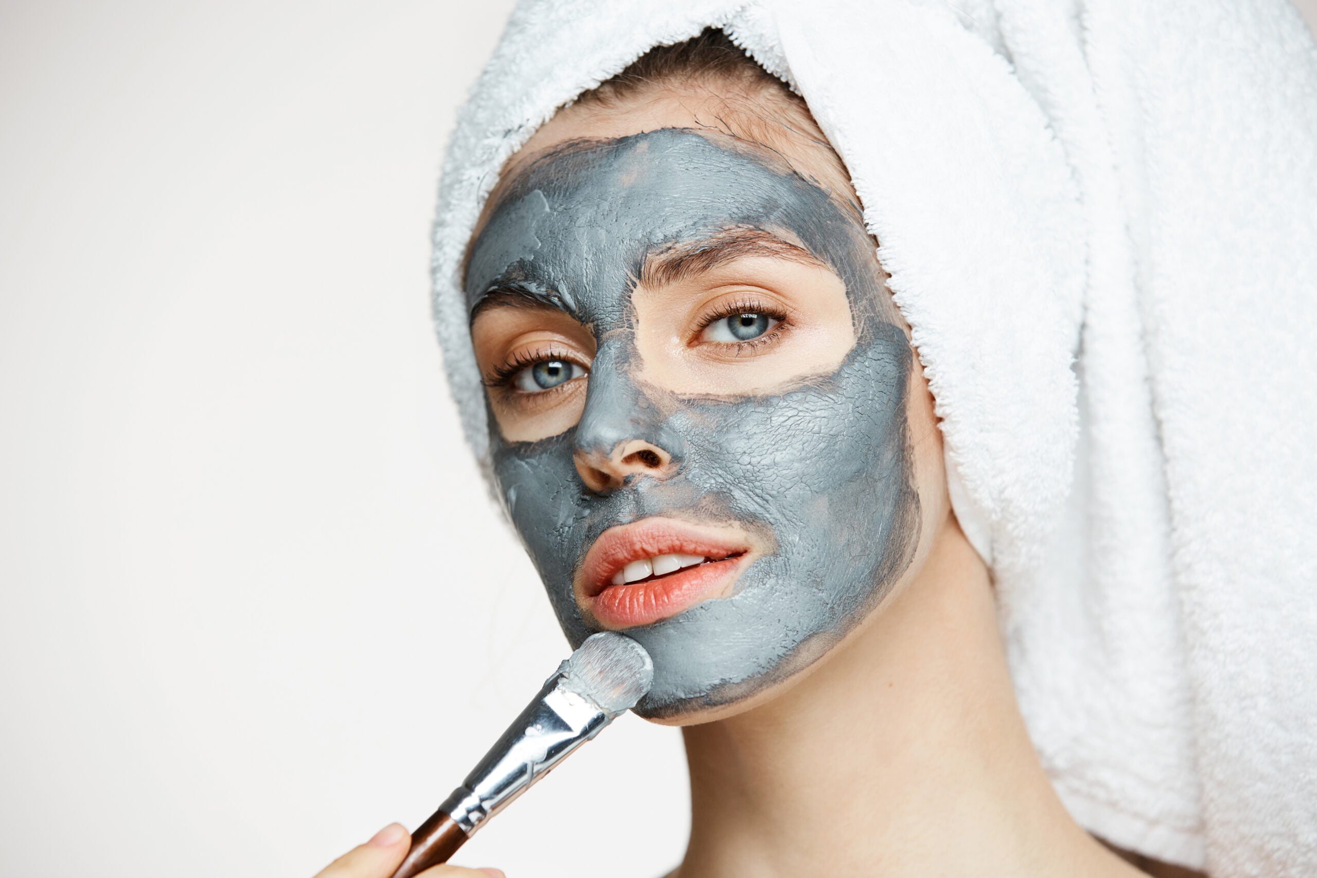 Young beautiful girl in towel on head covering face with mask smiling looking at camera over white background. Beauty cosmetology and spa. Copy space.